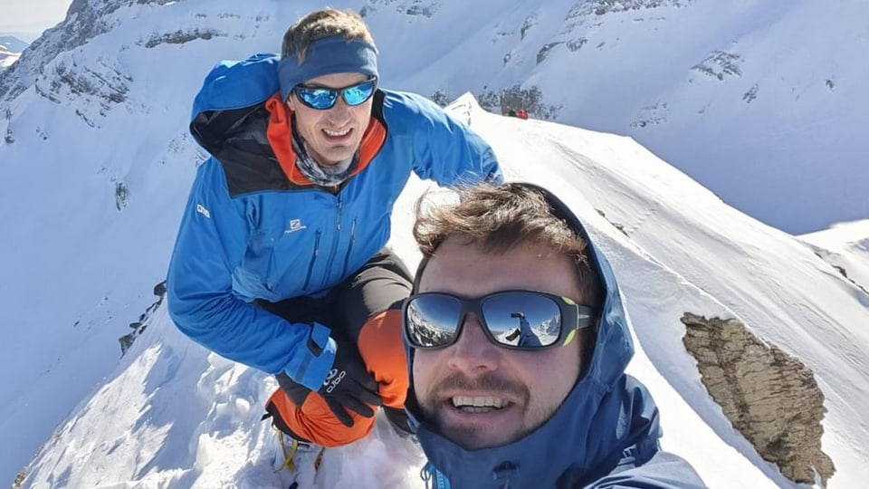 Zwei Männer in Windjacken und Sonnenbrillen stehen auf einem verschneiten Berggipfel.