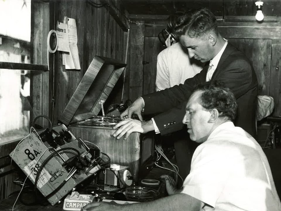 Die Schwarz-Weiss-Fotografie zeigt drei Männer, die vor technischen Geräten in einem Zimmer stehen oder sitzen.