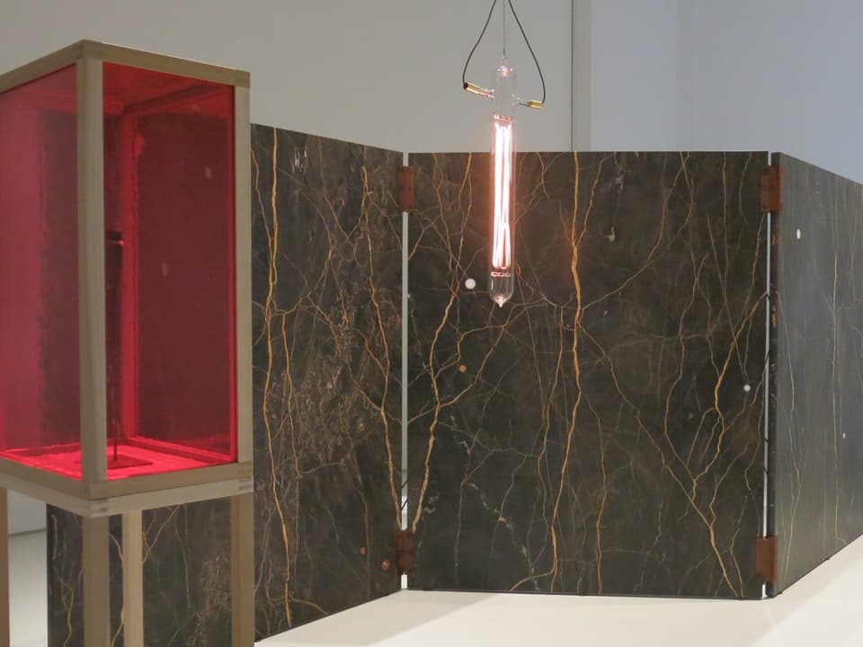 Eine längliche Glühbirne neben rotem Glaskasten, vor marmorartiger Wand 