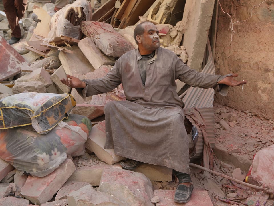 Ein Mann sitzt inmitten eines Trümmerhaufens und hebt die Hände gen Himmel.