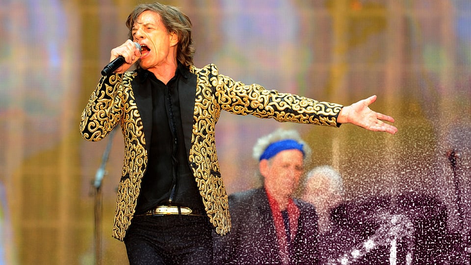 Mick Jagger trägt ein goldenes Jackett und sing in sein Mikrophon. 