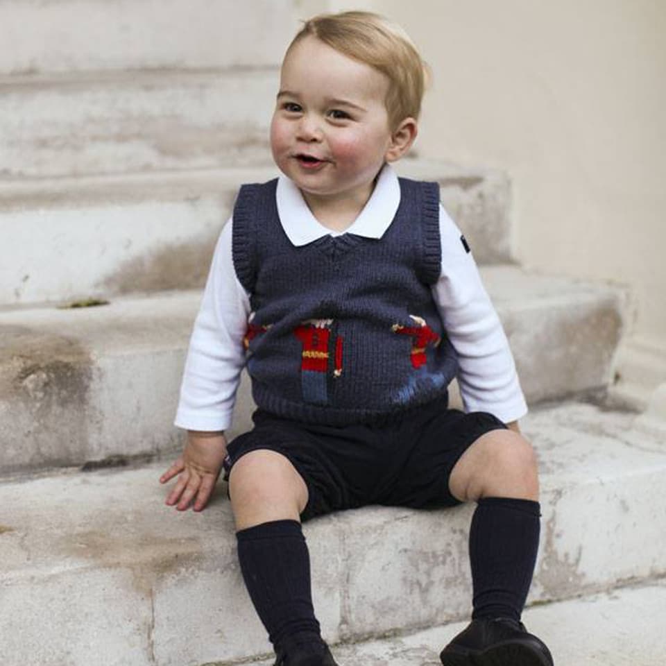 Prinz George in kurzen Hosen auf einer Steintreppe sitzend und lachend.