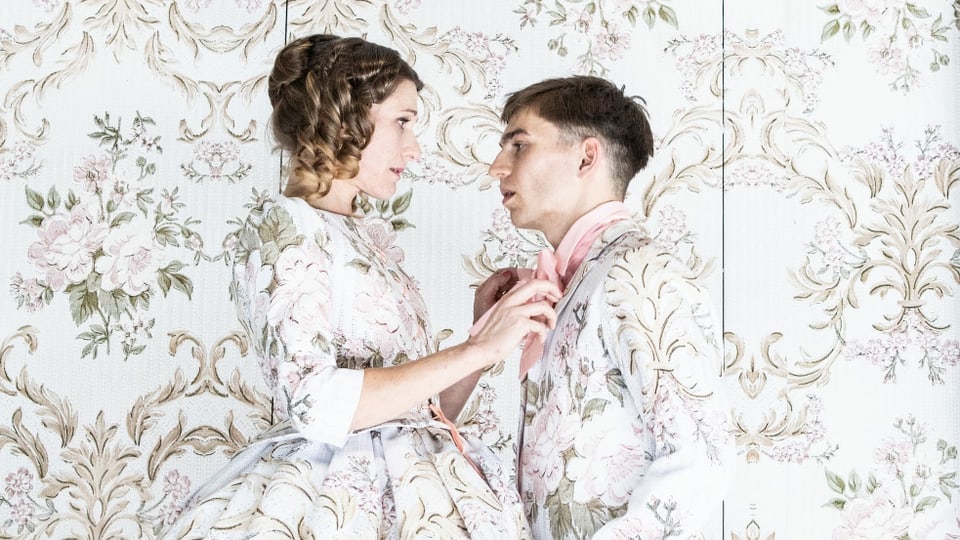 Theaterszene: Ein Paar in Kleidung mit Blumenmustern. Die Tapate im Hintergrund hat das gleiche Muster.
