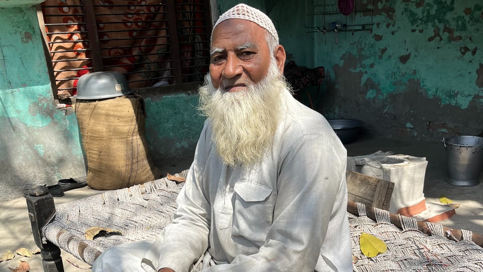 Älterer Mann mit weissem Bart und traditioneller Kleidung sitzt vor einem Haus.
