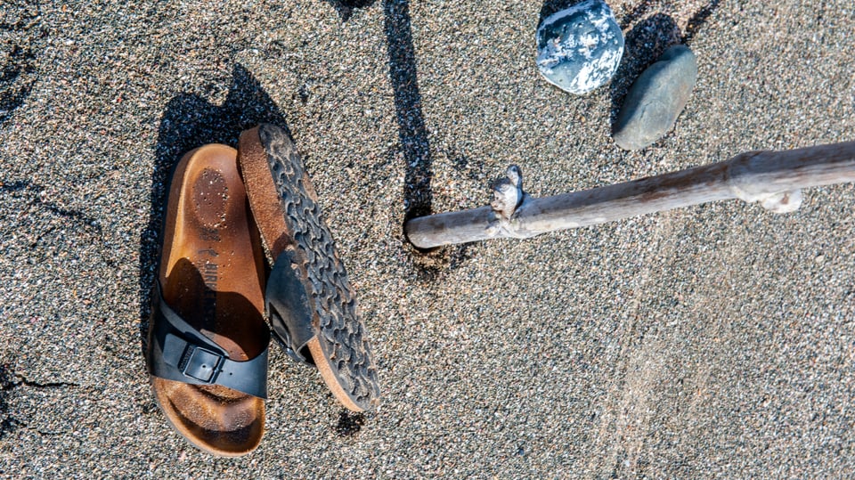 Zwei Sandalen mit einem schwarzen Lederriemen und einer Schnalle liegen auf Sand. 