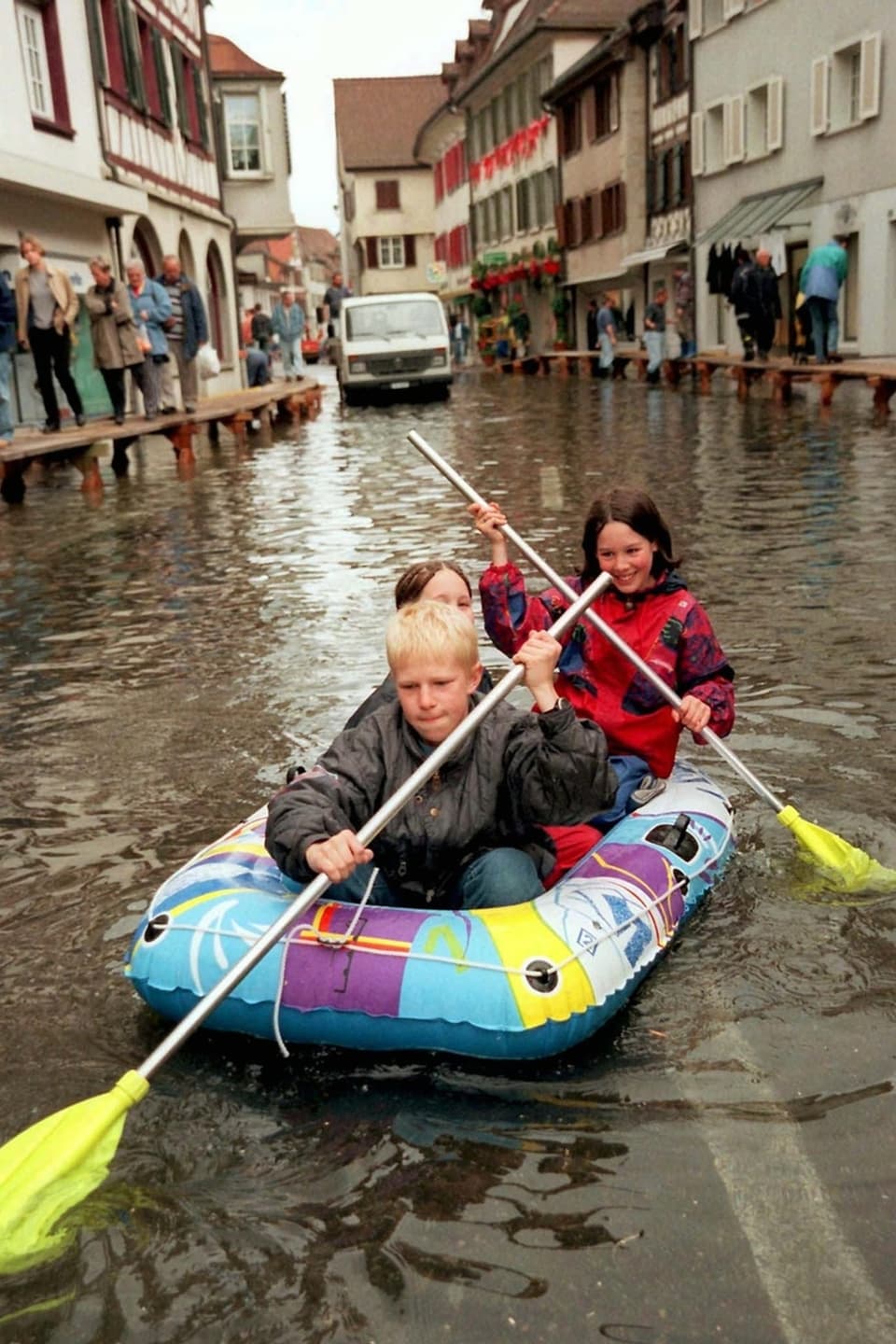 Zwei Kinder paddeln in einem Schlauchboot auf einer überfluteten Strasse in einer Stadt.