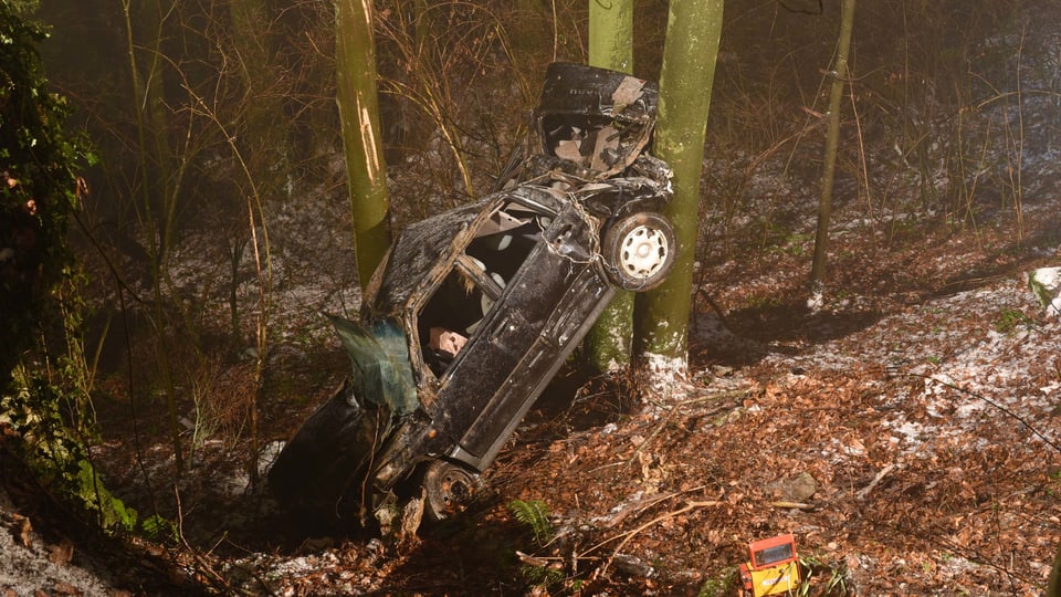 Ein total zerstörtes Auto in einem steilen Waldstück.