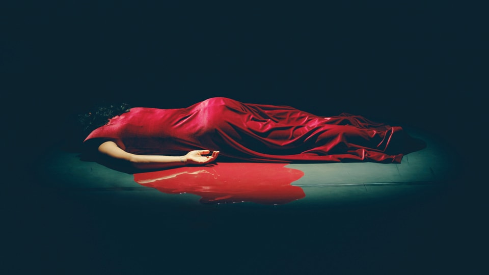 Eine Frau in rotem Kleid liegt auf der Bühne. Unter und neben ihr zeigt sich ein roter Fleck.