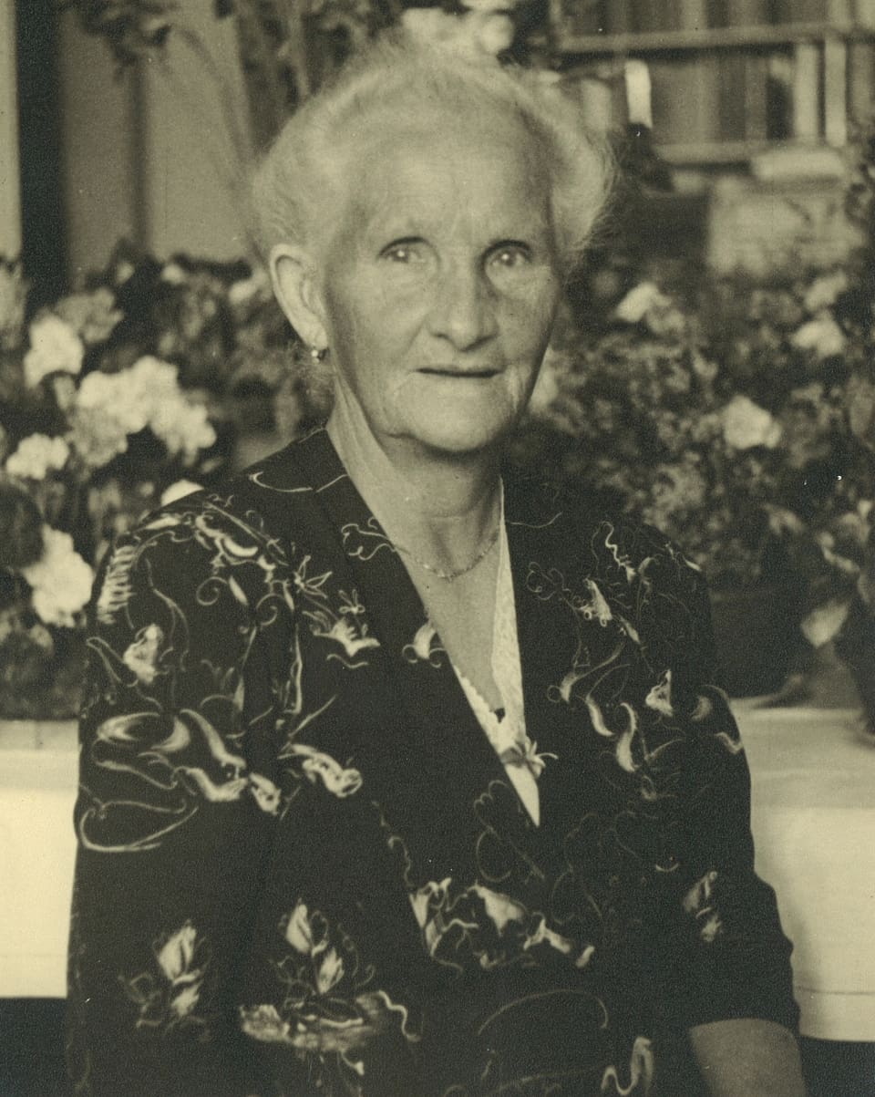 Schwarz-Weiss-Fotografie mit dem Porträt einer weisshaarigen Frau, die vor Blumen sitzt.