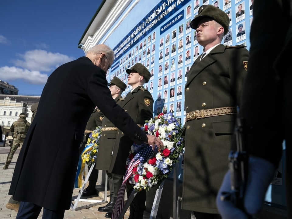 Mann in schwarzem Mantel legt rot-weissen Blumenkranz nieder, vor einer blauen Wand mit Fotos. Daneben stehen Soldaten in Uniform.