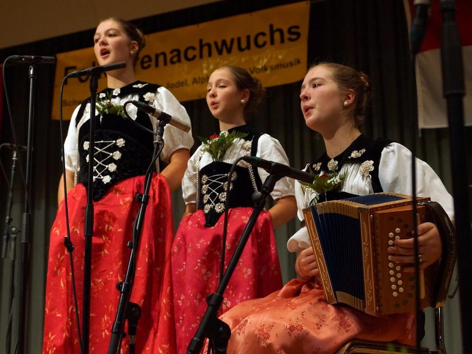 Zwei junge Sängerinnen singen stehend und werden von der Schwester am Schwyzerörgeli begleitet.