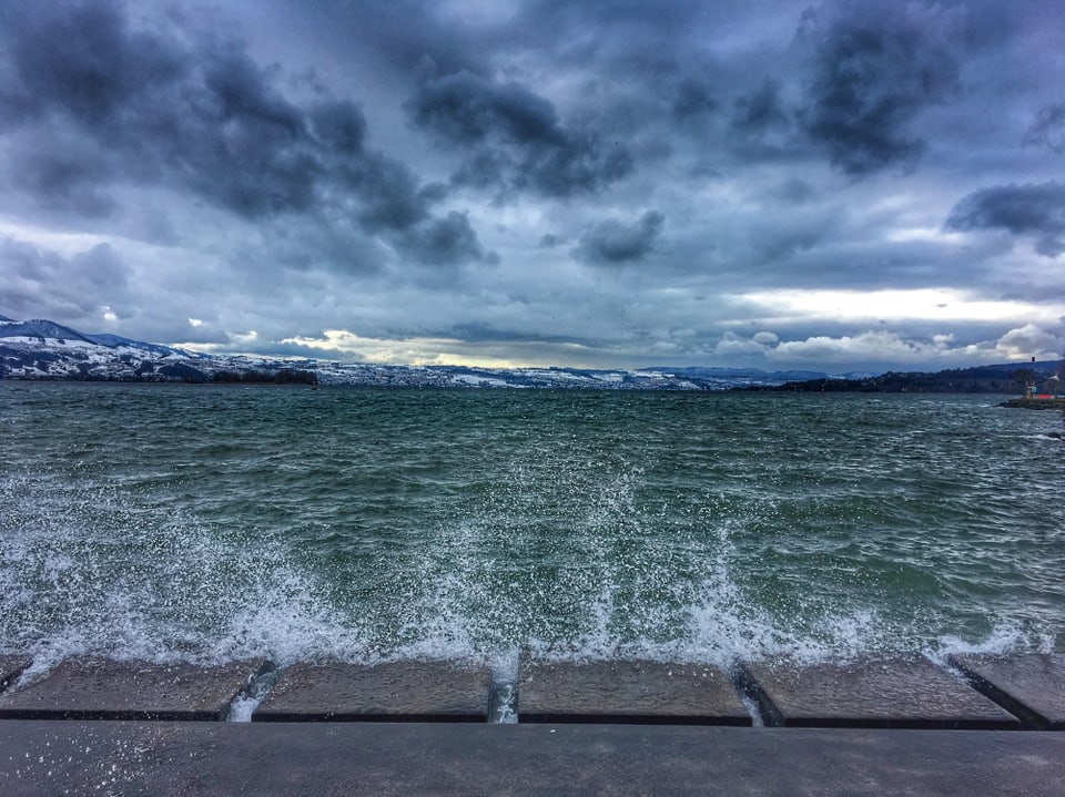 Blick auf den Zürichsee. Der See ist wellig und das Wasser spritzt ans Ufer. Der Himmel wirkt mit dunklen Wolken bedrohlich.
