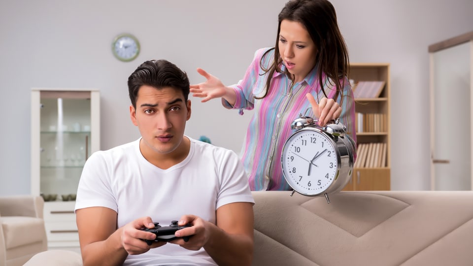 Ein Mann sitzt auf dem Sofa und spielt ein Konsolen-Game, eine Frau erinnert ihn mit einem überdimensionierten Wecker daran, wie viel Zeit er damit schon verbracht hat.