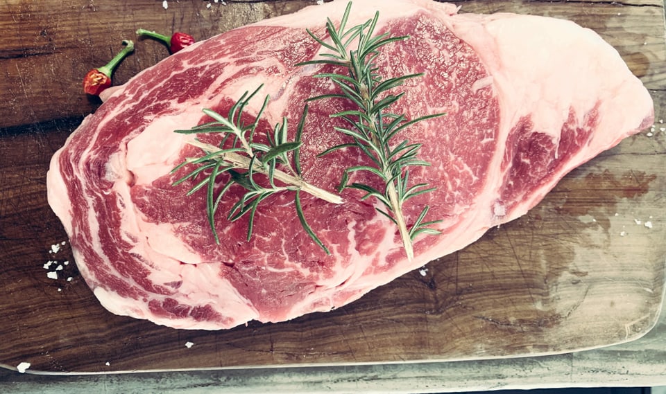 Ein rohes gesalzenes Steak mit frischem Rosmarin und zwei Cihilischoten auf einem Holzbrett.