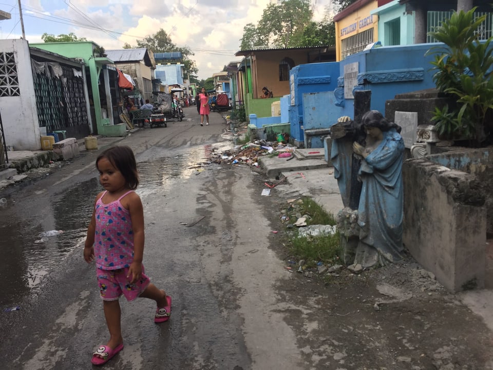 Ein kleines Mädchen überquert die regennasse Strasse, die von Mausoleen gesäumt ist. Im Hintergrund eine Engelsskulptur, die grösser als das Mädchen ist.