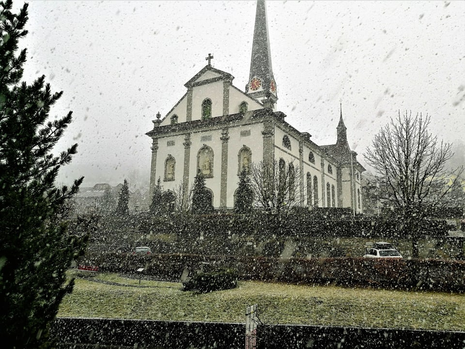Vor einer Kirche fallen dicke Schneeflocken, der Park davor ist noch grün.