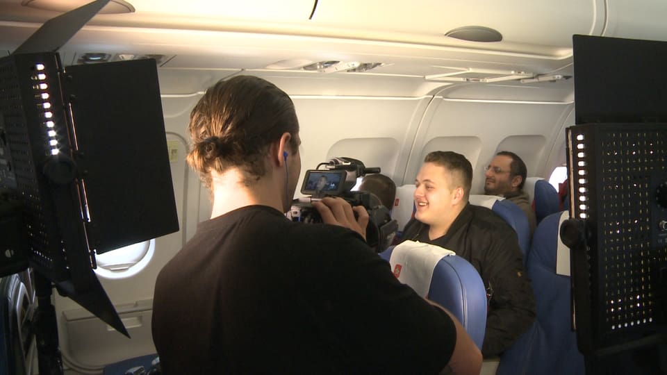Ein junger Mann sitzt im Flugzeug. Er ist von Kameras umgeben, die einen Werbespot drehen.