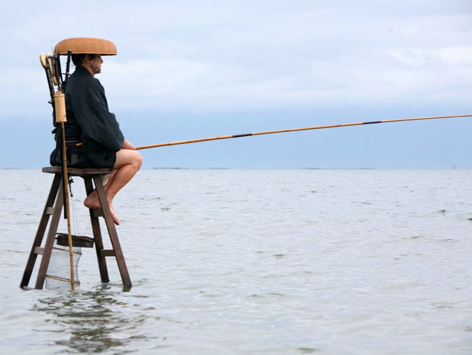 Ein Mann sitzt auf einem hohen Holzstuhl mitten im Wasser und hält eine lange Fischerrute.
