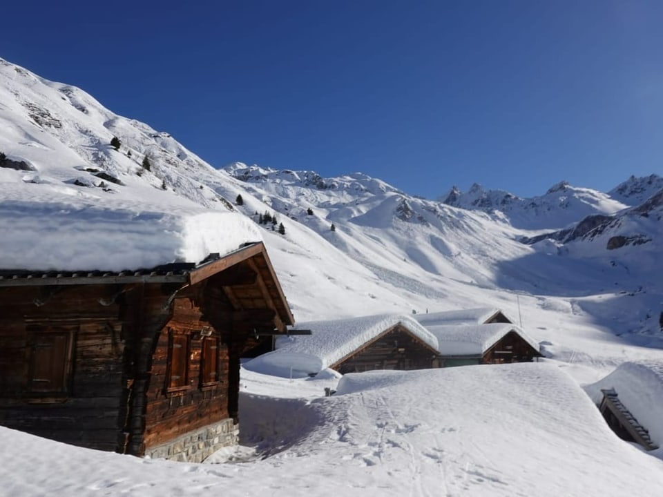 Berghütten mit Schnee im Vordergrund. Blauer Himmel.
