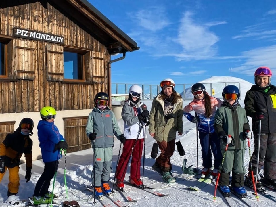 Eine Gruppe Skifahrer:innen und Snowboarder:innen steht auf einem Gipfel vor einem Haus