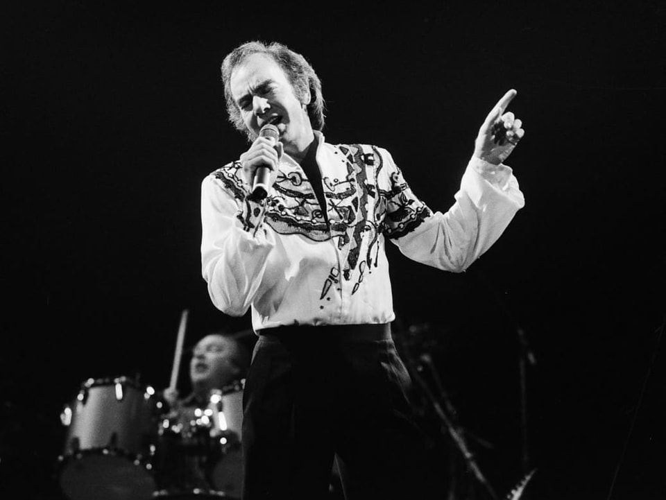 Schwarz-Weiss-Aufnahme von Neil Diamond, der mit einem Mikrofon in der Hand singt. Im Hintergrund sitzt ein Mann am Schlagzeug. 