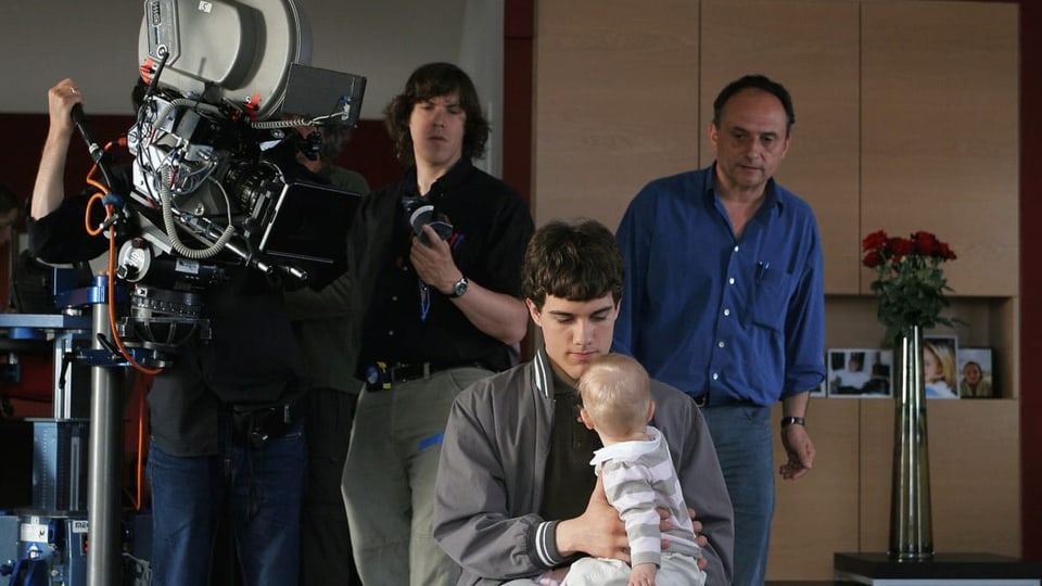 Filmset: Im Vordergrund ein junger Mann mit Baby auf dem Schoss. Im Hintergrund drei Männer und eine grosse Kamera.