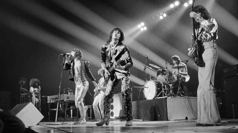 Mick Jagger singt in Richtung Kamera, die Band spielt. Sie tragen bunte Kleidung.