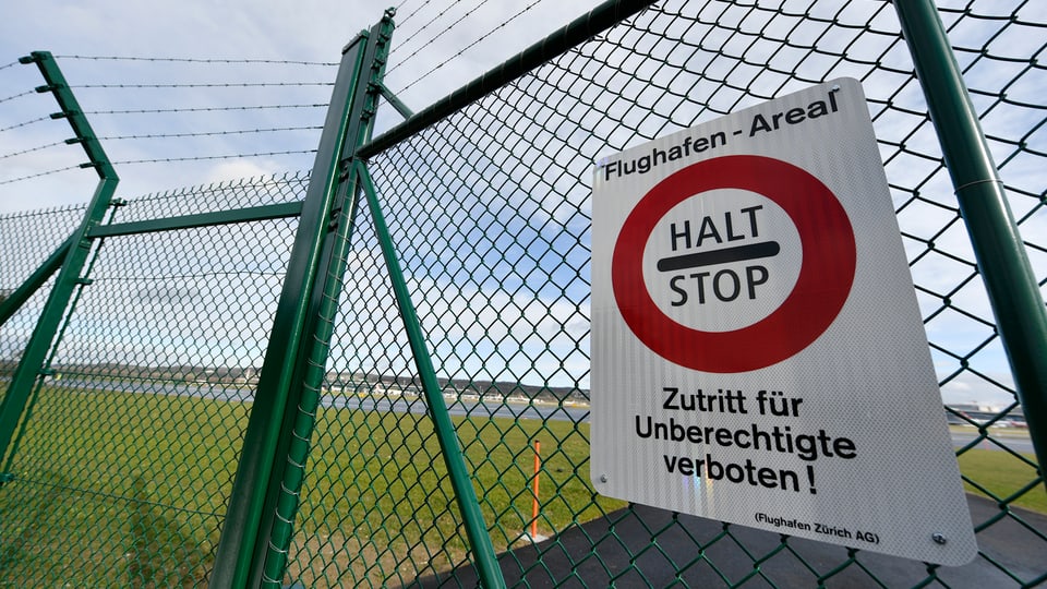Ein Schild hängt an einem Zaun, darauf steht: "Halt - Stop". Dahinter ist eine Landebahn am Zürcher Flughafen.