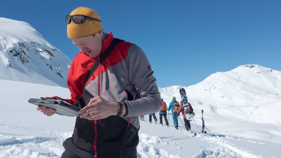 Ein Mann steht mit einem Tablet in einer Schneelandschaft in den Bergen. Hinter ihm sind Wintersportler sichtbar, die an einer Kamera vorbeigehen. 