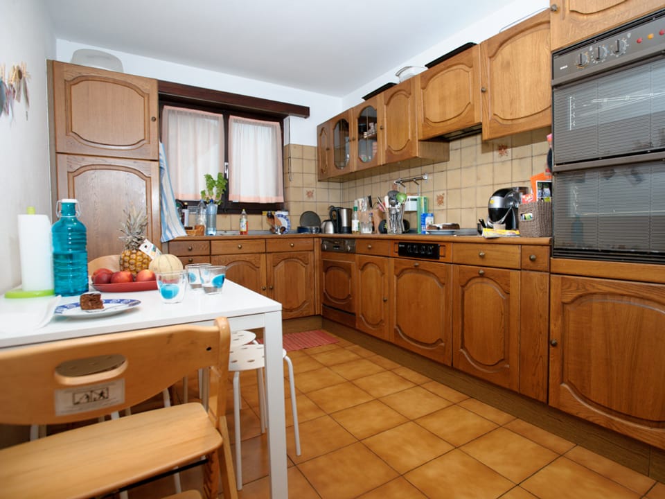 Blick in die Küche. Die Küchenkombination ist in eher dunklem Holz gehalten. Links steht ein kleiner weisser Küchentisch.