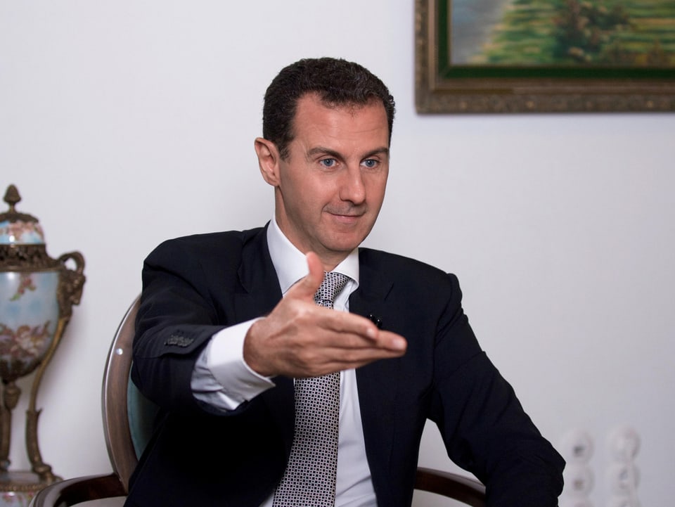 Bashar Al-Assad sitzt auf einem Stuhl und streckt die Hand aus