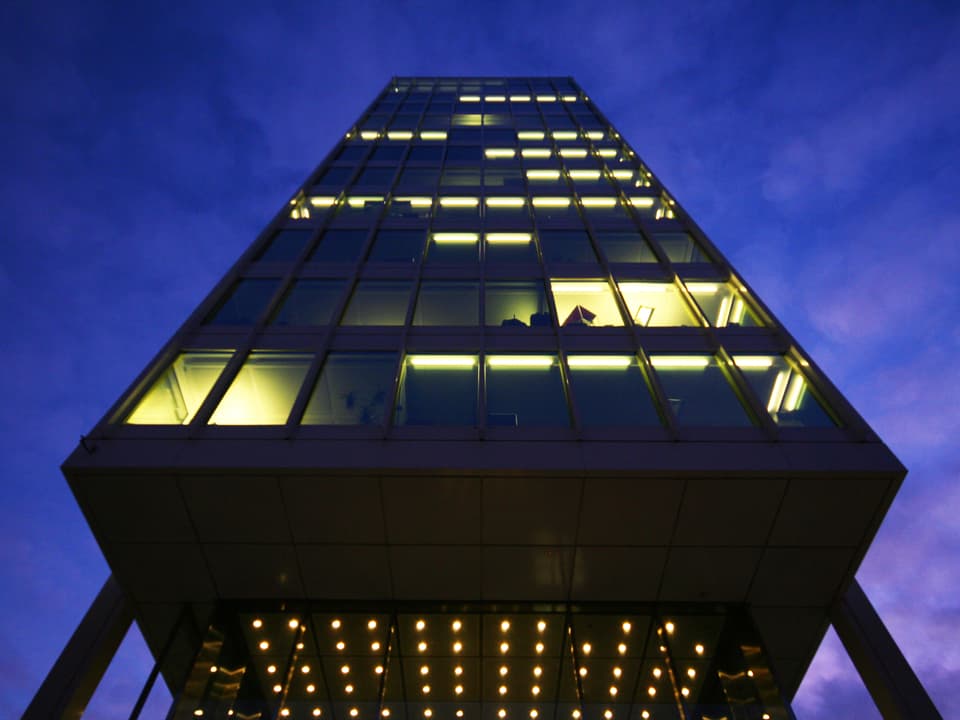 Ein modernes Gebäude steht auf Pfeilern, aufgenommen aus der Froschperspektive. Aus den Fenstern scheint Licht. 