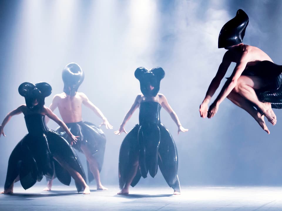 Drei Figuren mit schwarzer Maske stehen auf der Bühne, rechts davon springt eine vierte Figur hoch.