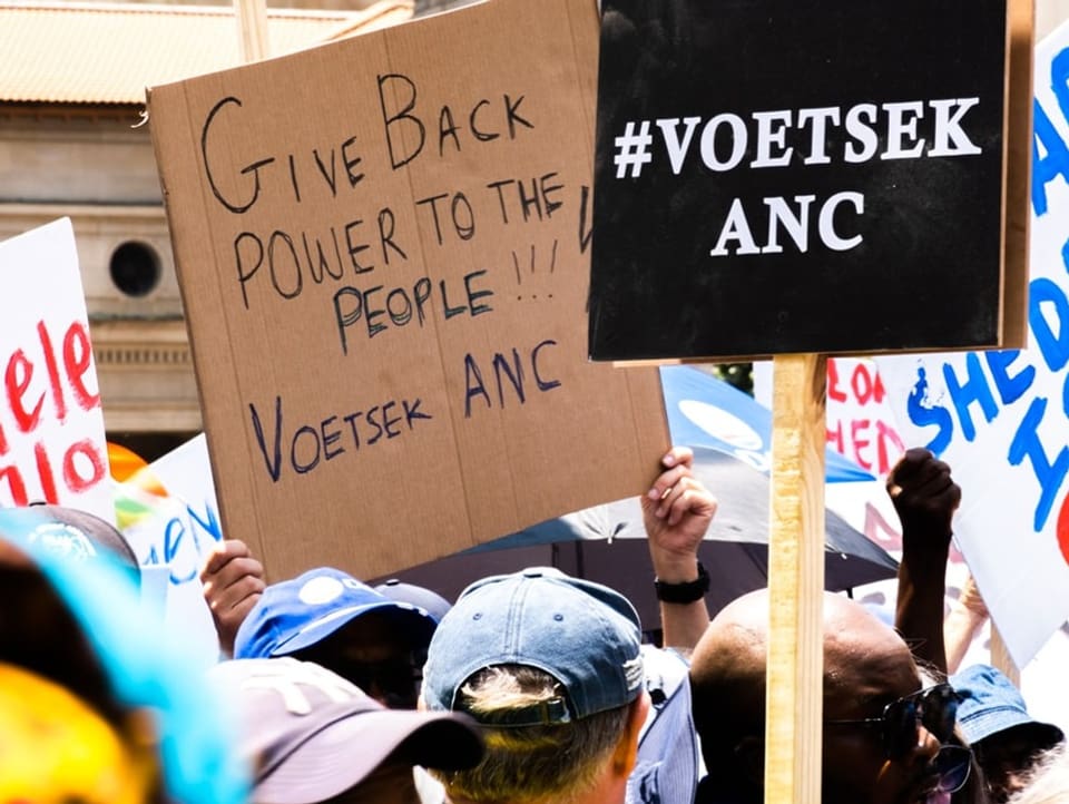 Nahaufnahme von Demonstrantinnen und Demonstranten. Auf Plakaten steht «Voetsek ANC»