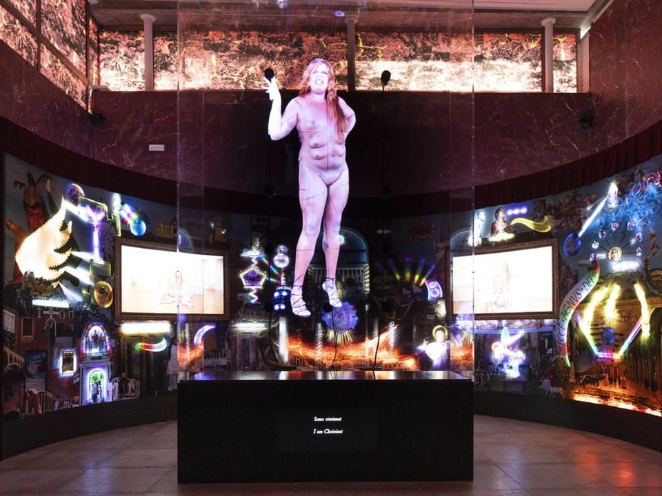 Holographische Darstellung einer stehenden Person umgeben von digitalen Bildschirmen in einer Ausstellung.