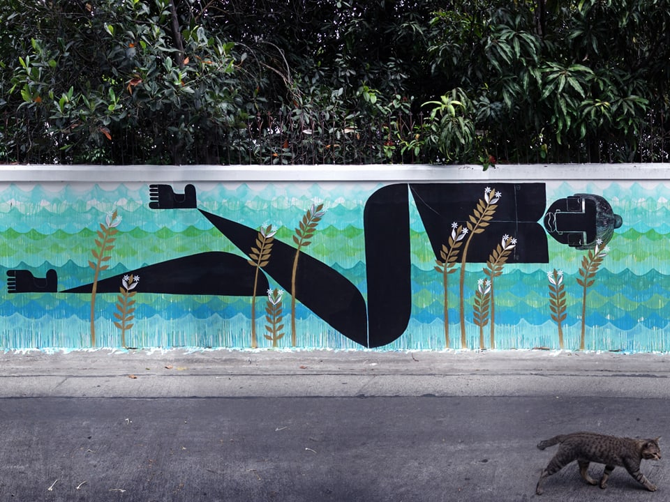 Eine Wandmalerei auf einer Mauer, die eine weibliche Figur am Schwimmen zeigt. Im Vordergrund geht eine Katze vorbei. 
