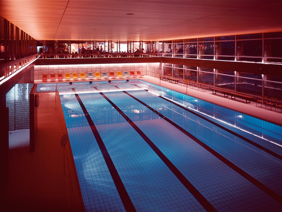 Indoor-Swimmingpool, am Ende der Schwimmbahnen stehen farbige Liegestühle.