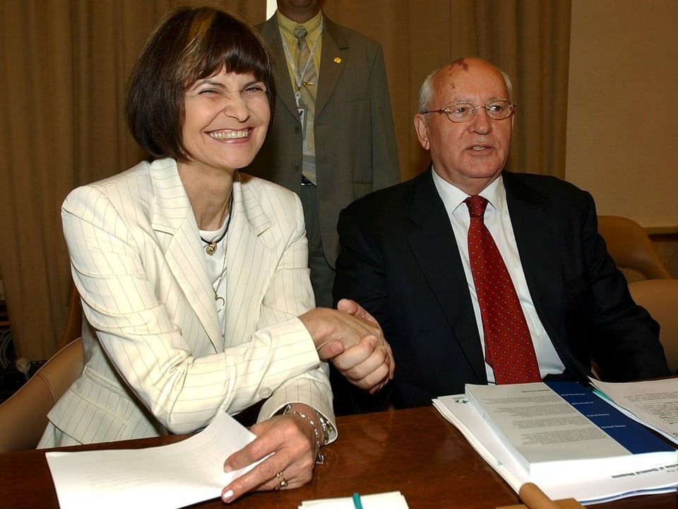 Micheline Calmy-Rey sitzt neben Gorbatschow und lächelt. Sie schütteln sich die Hand.