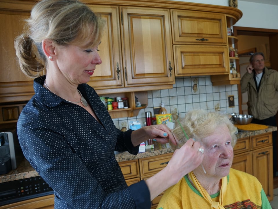 Eine Frau kämmt die Haare einer alten Frau, die in der Küche sitzt.