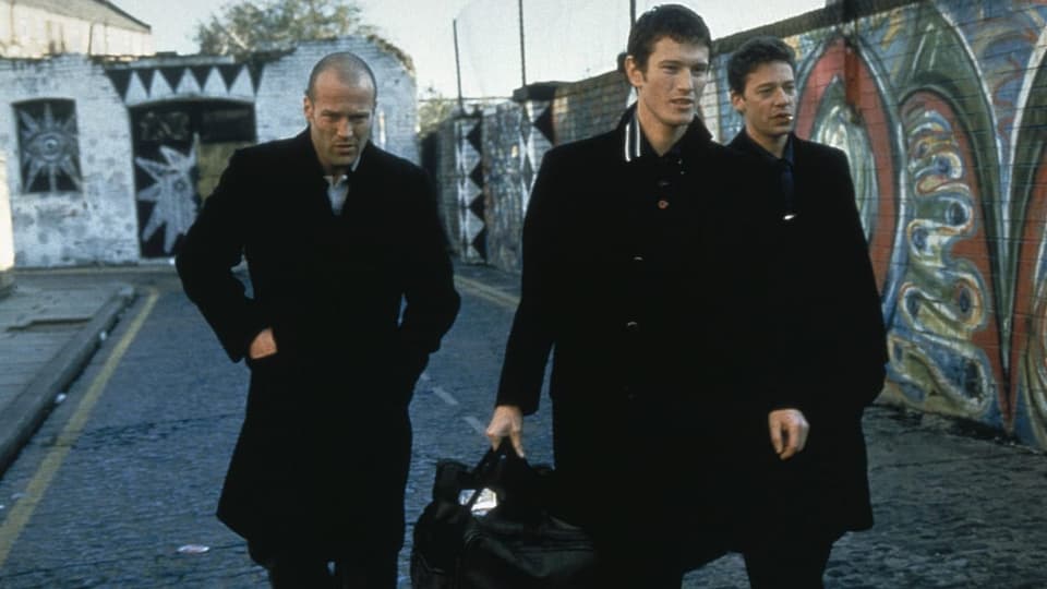 Foto von drei jungen Männern in schwarz, laufen auf Strasse mit Graffiti-Mauer, Mann links mit Glatze, grimmig.
