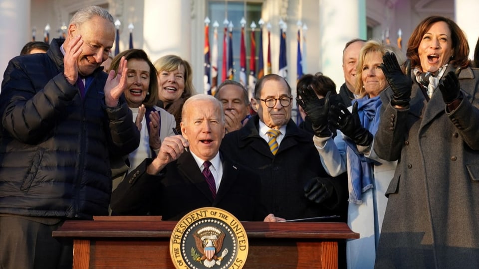 US-Präsident Joe Biden, umgeben von applaudierenden Politikern vor dem Weissen Haus, unterschreibt etwas an einem Tisch.