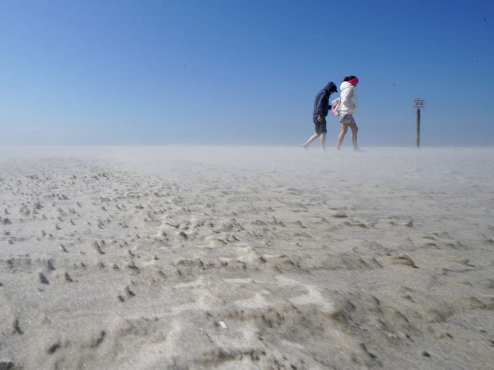 Zwei Personen laufen im Sandsturm am Strand.