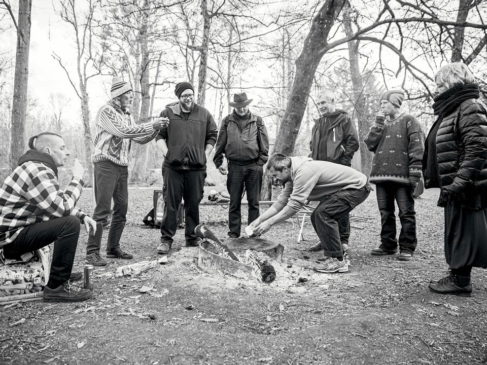 Schwarzweissbild von 7 Menschen, die in einem Wald um eine Feuerstelle stehen.