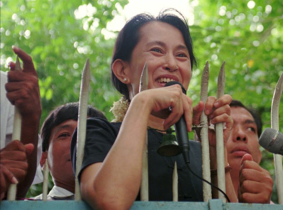Aung San Suu Kye steht hinter Gitterstäben und sprich in ein Mikrophon. 