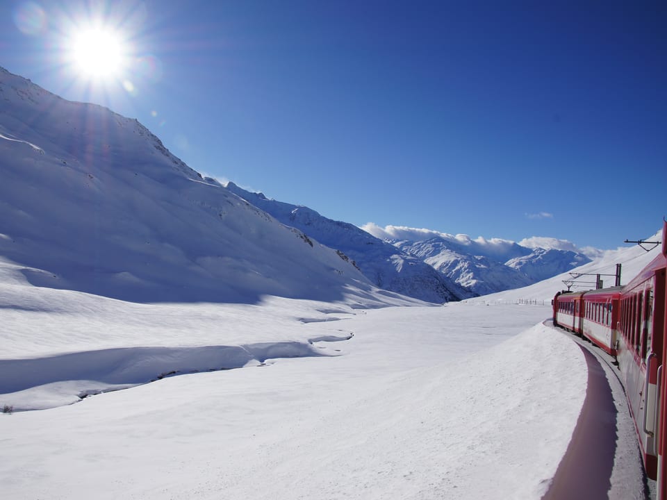 Blick aus dem Zug über eine verschneite Berglandschaft. Die Sonne scheint vom blauen Himmel.