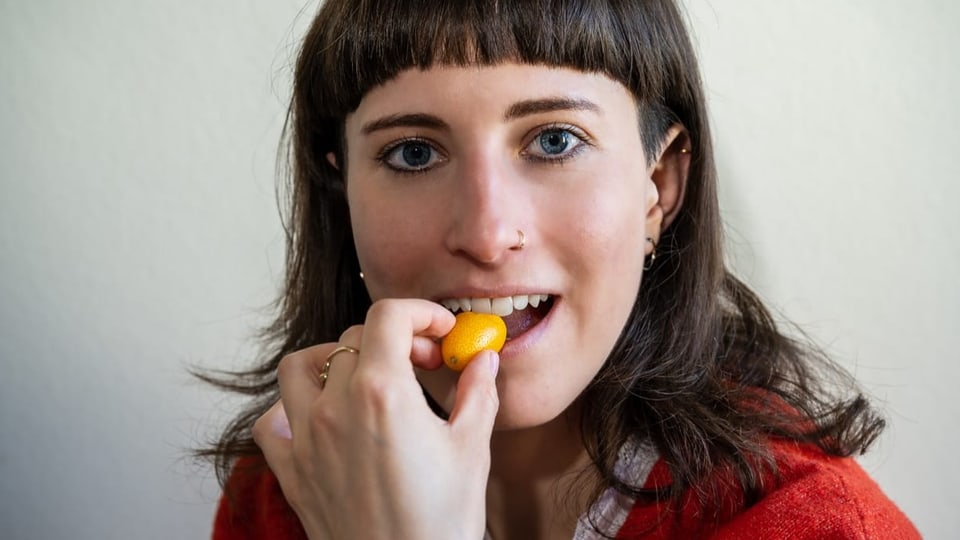 Junge Frau mit dunklen Haaren und blauen Augen blickt in Kamera, beisst auf kleines, orangenes Früchtchen.