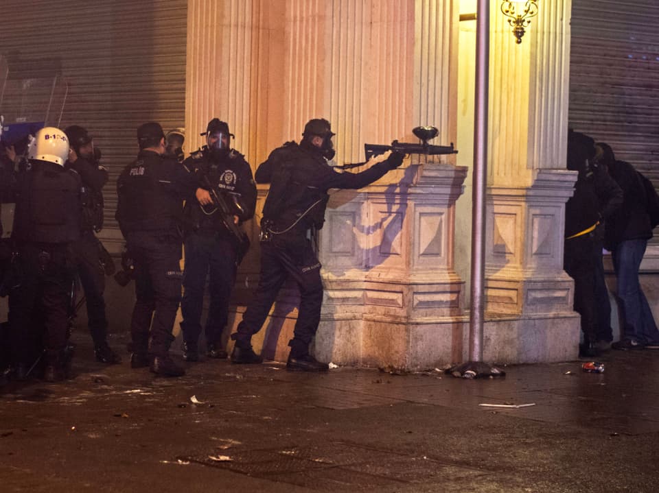Polizisten mit Gasmasken stehen hinter einem Ecken, einer richtet seine Waffe auf jemanden.