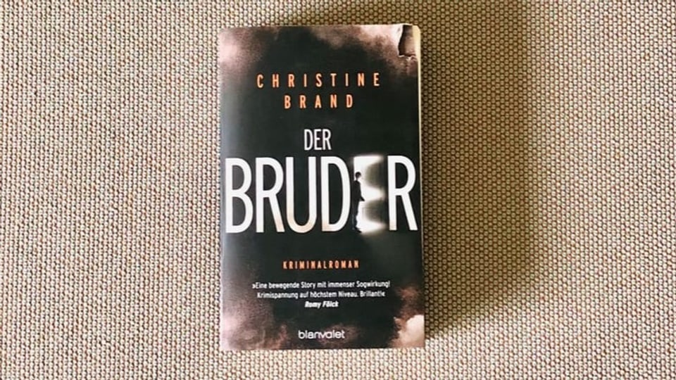 «Der Bruder» von Christine Brand liegt auf einem beigen Sofastoff