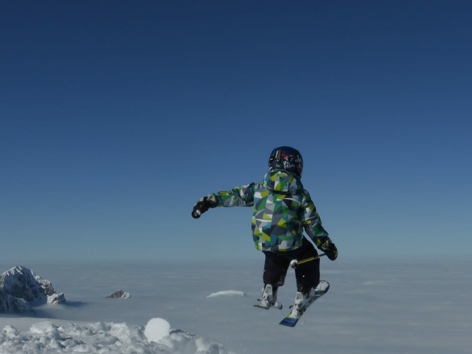 Ein Kind springt mit den Ski's. Im Hintergrund liegt ein Nebelmeer.