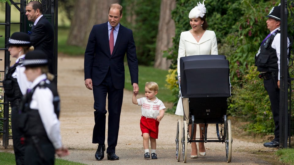 Prinz George geht an der Hand von Prinz William. Neben ihnen schiebt Kate einen Kinderwagen.